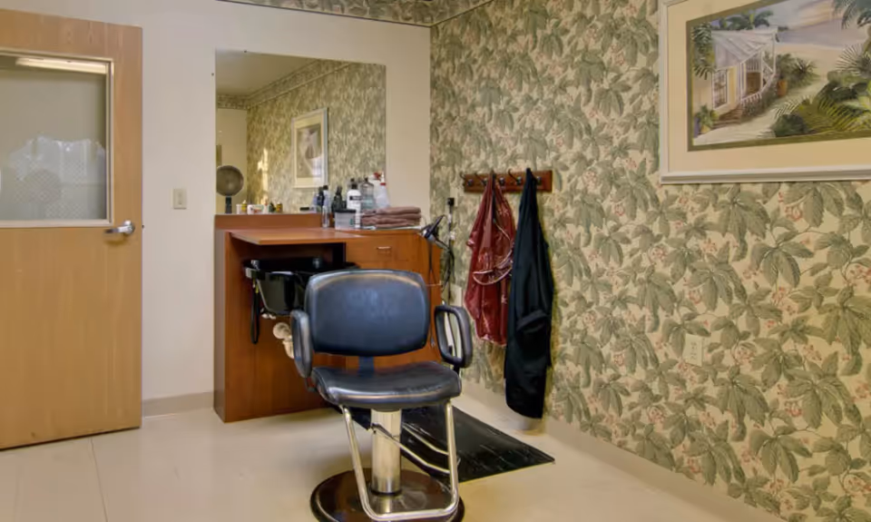 Interior room with a salon/barber chair in front of a sink and mirror, floral wallpaper, coats hanging and a framed painting.