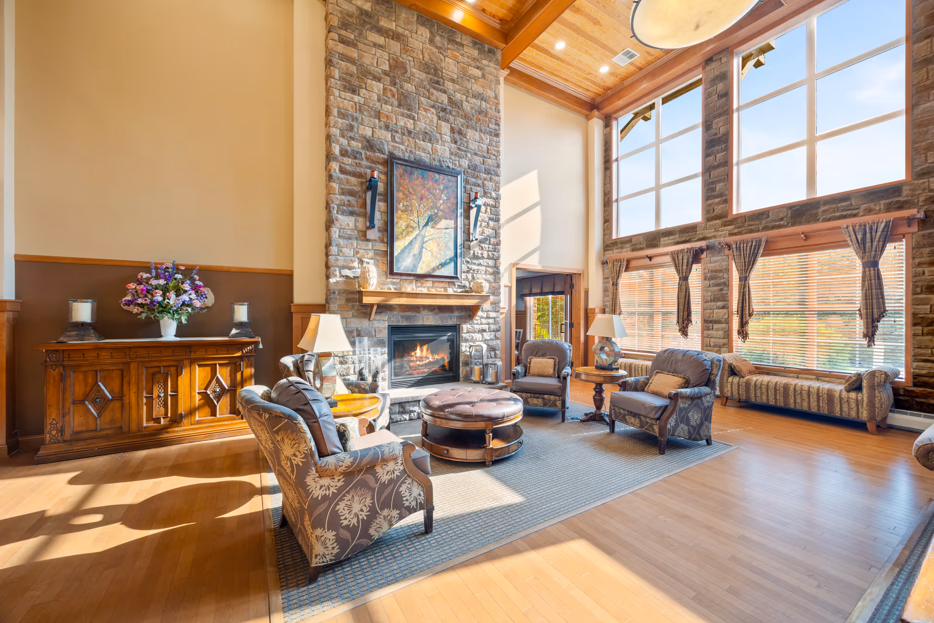 A spacious living room with high wooden ceilings and large windows letting in natural light. The room features a stone fireplace with a painting above it, surrounded by four upholstered armchairs and a round ottoman on a rug. There is a wooden sideboard with a vase of flowers and two candles, and a cushioned bench along the window wall.