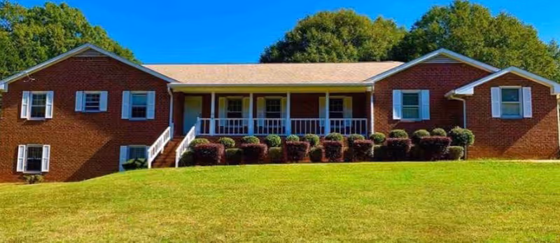Front exterior view of a single-story brick building with white trim and green shutters, featuring a covered porch with white railings and steps leading up to it, surrounded by neatly trimmed bushes and a grassy lawn under a clear blue sky.
