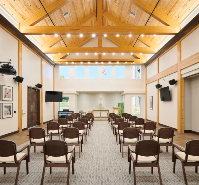 Interior view of a chapel or meeting room with rows of wooden chairs with white cushions arranged facing an altar at the front. The room features a high wooden vaulted ceiling with exposed beams and multiple windows letting in natural light. There are two wall-mounted televisions on either side and framed pictures on the left wall.