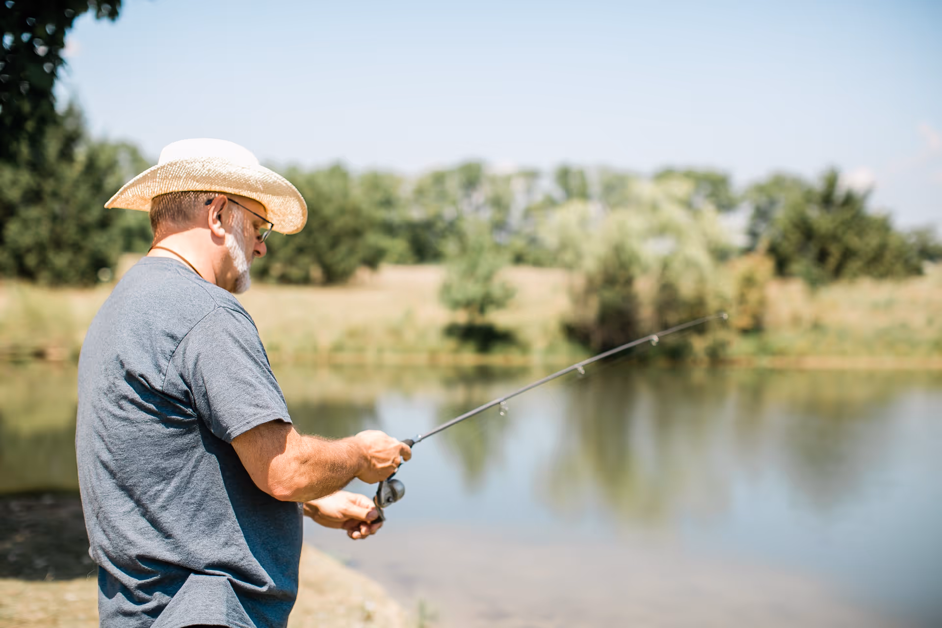 An elderly man wearing a straw hat and glasses is fishing by a calm pond surrounded by greenery on a sunny day.