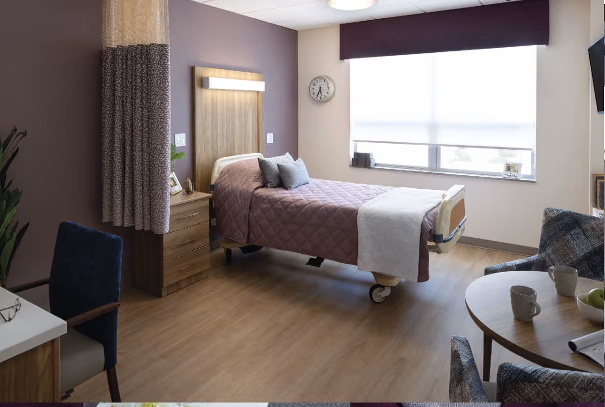A well-lit bedroom in a senior living facility with a single bed covered in a purple quilt and a white blanket at the foot. The room features a wooden nightstand with decorative items, a clock on the wall, a large window with a purple valance, a round table with two chairs, and a plant in the corner.