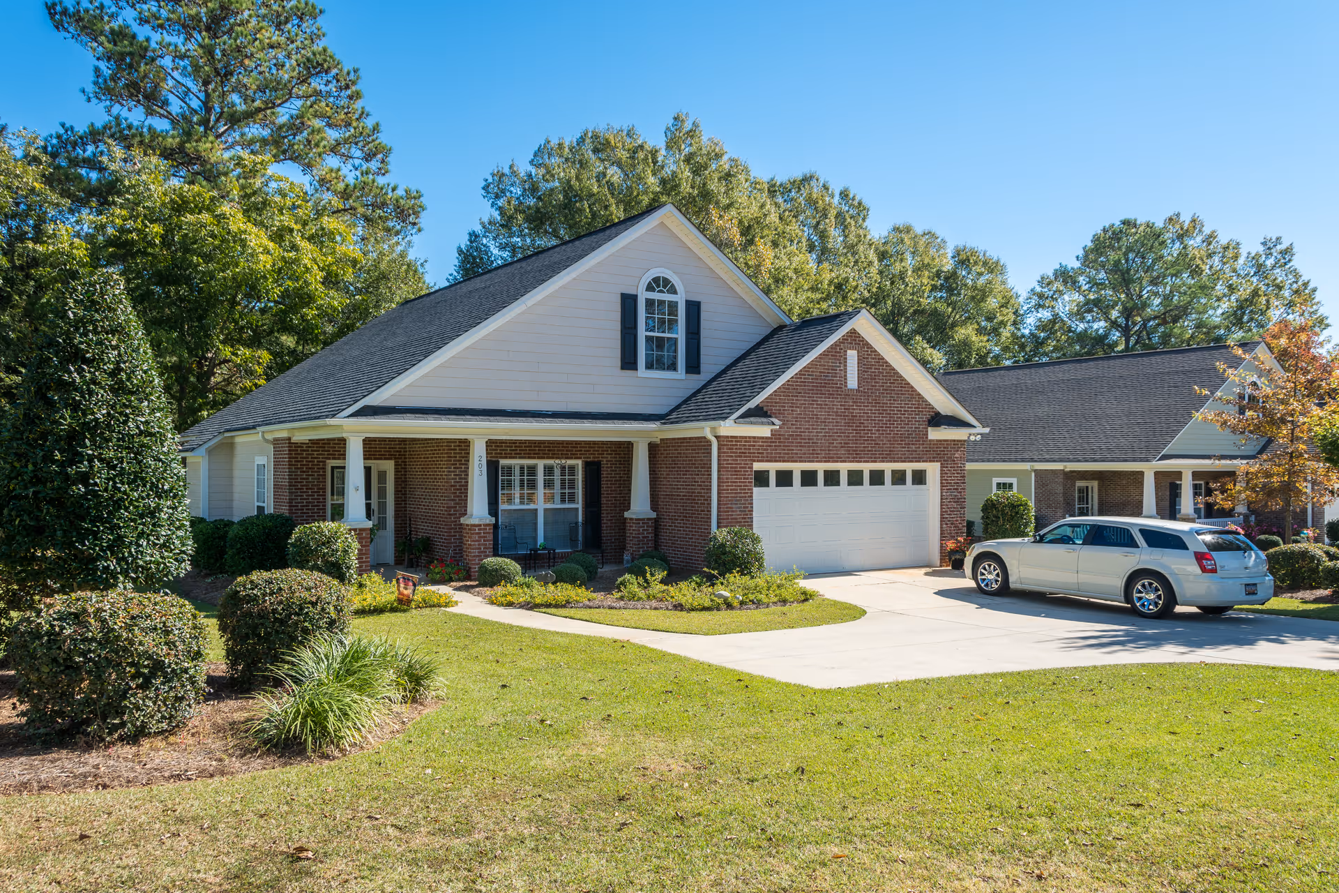 A single-story brick and siding house with a front porch, a two-car garage, and a driveway with a silver car parked on it. The house is surrounded by neatly trimmed bushes, a green lawn, and tall trees under a clear blue sky.