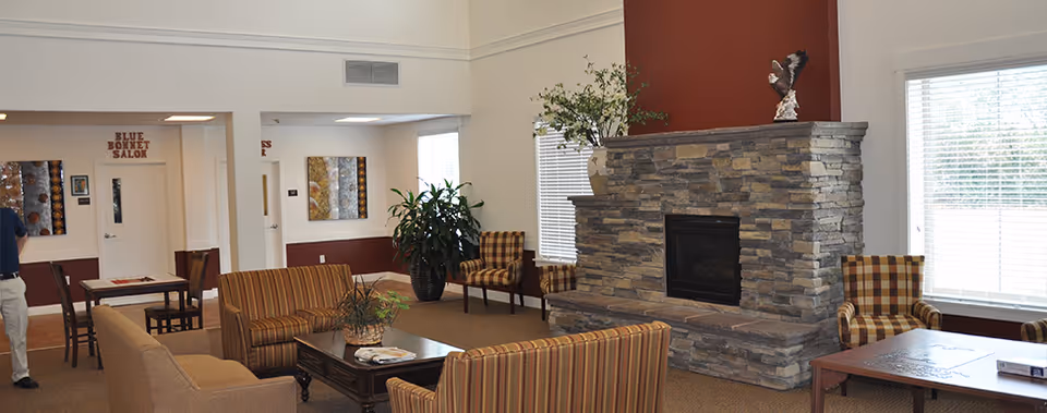 A cozy assisted living facility common area with a stone fireplace as the focal point. The room features striped and plaid upholstered chairs and sofas arranged around wooden coffee tables. There are large windows with blinds allowing natural light to fill the space, and decorative plants and artwork adorn the walls. A sign reading 'Blue Bonnet Salon' is visible above a doorway in the background.