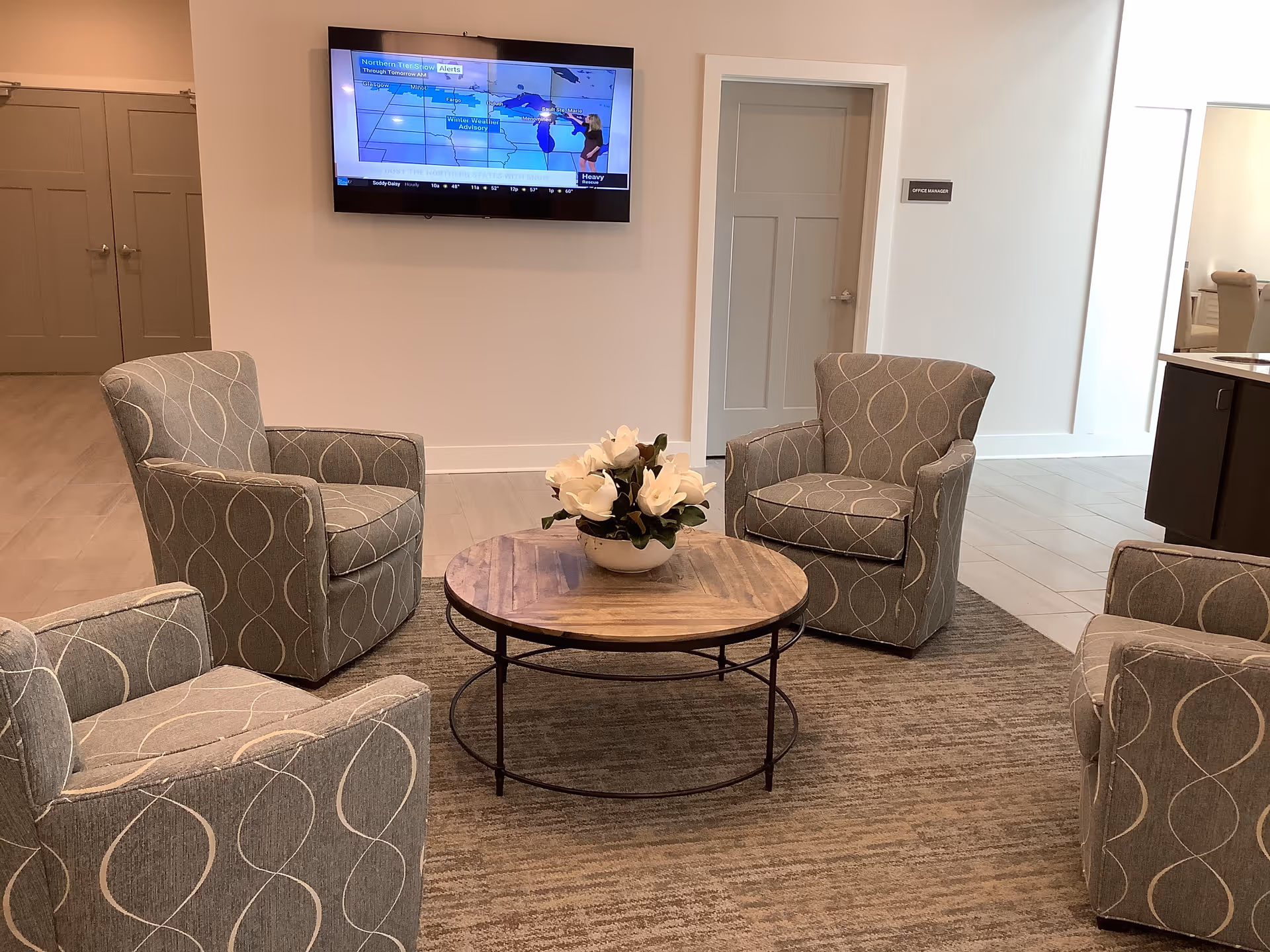 A cozy seating area with four gray armchairs arranged around a round wooden coffee table with a flower arrangement on top. A flat-screen TV is mounted on the wall displaying a weather forecast. There are two closed doors and a sign labeled 'Office Manager' on the wall in the background.