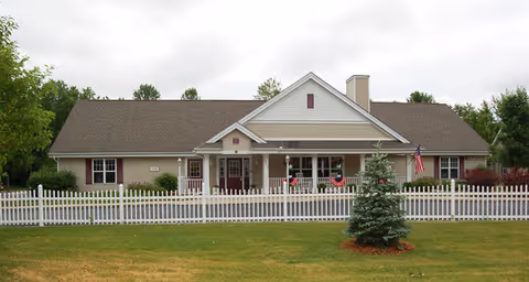 Single-story residential-style building with a white picket fence in front, a small evergreen tree on the lawn, and an American flag hanging near the entrance under a covered porch.