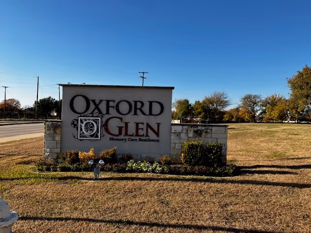 Outdoor view of a stone and concrete sign for Oxford Glen Memory Care Residence, situated on a grassy area with some bushes and trees in the background under a clear blue sky.