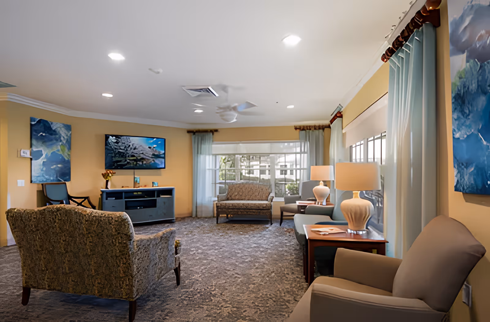 A cozy living room area in a senior living facility with multiple upholstered chairs and sofas arranged around a carpeted floor. There is a flat-screen TV mounted on a yellow wall, two table lamps on wooden side tables, large windows with light blue curtains allowing natural light, and abstract blue artwork on the walls.