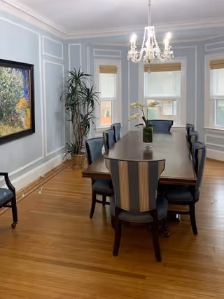 A formal dining room with a long wooden table surrounded by eight upholstered chairs. The room has light blue walls with white trim, three windows with beige blinds, a chandelier hanging above the table, a large potted plant in the corner, and a framed painting on the left wall.