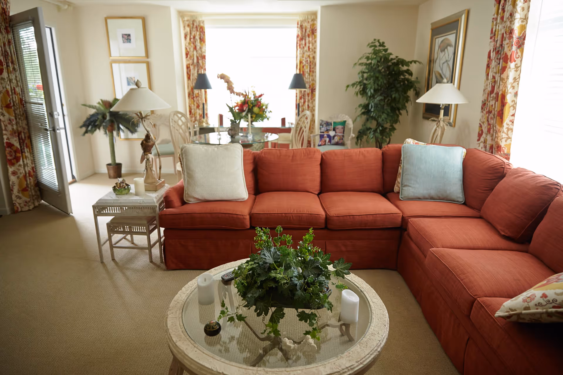 A cozy living room with a large red sectional sofa adorned with various cushions. In front of the sofa is a round glass coffee table with a green leafy plant and candles. Behind the sofa, there is a dining area with a glass table and chairs, floral curtains on the windows, and decorative lamps. The room is warmly lit with natural light coming through the windows.