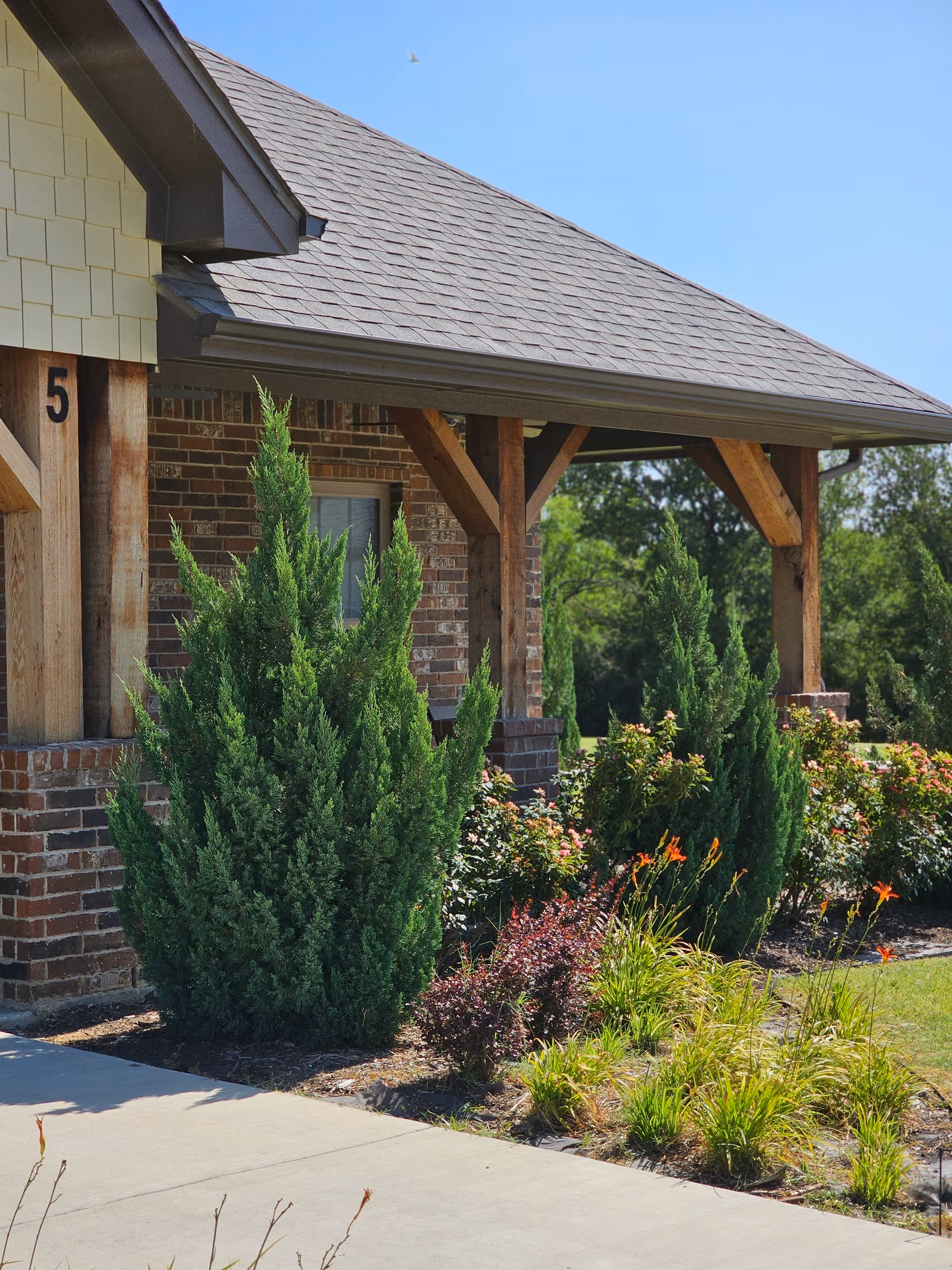 Exterior view of a brick building with wooden posts supporting a roof overhang. The building is surrounded by well-maintained landscaping including green shrubs, flowering plants, and a concrete walkway. The number 5 is visible on the building near the roof.