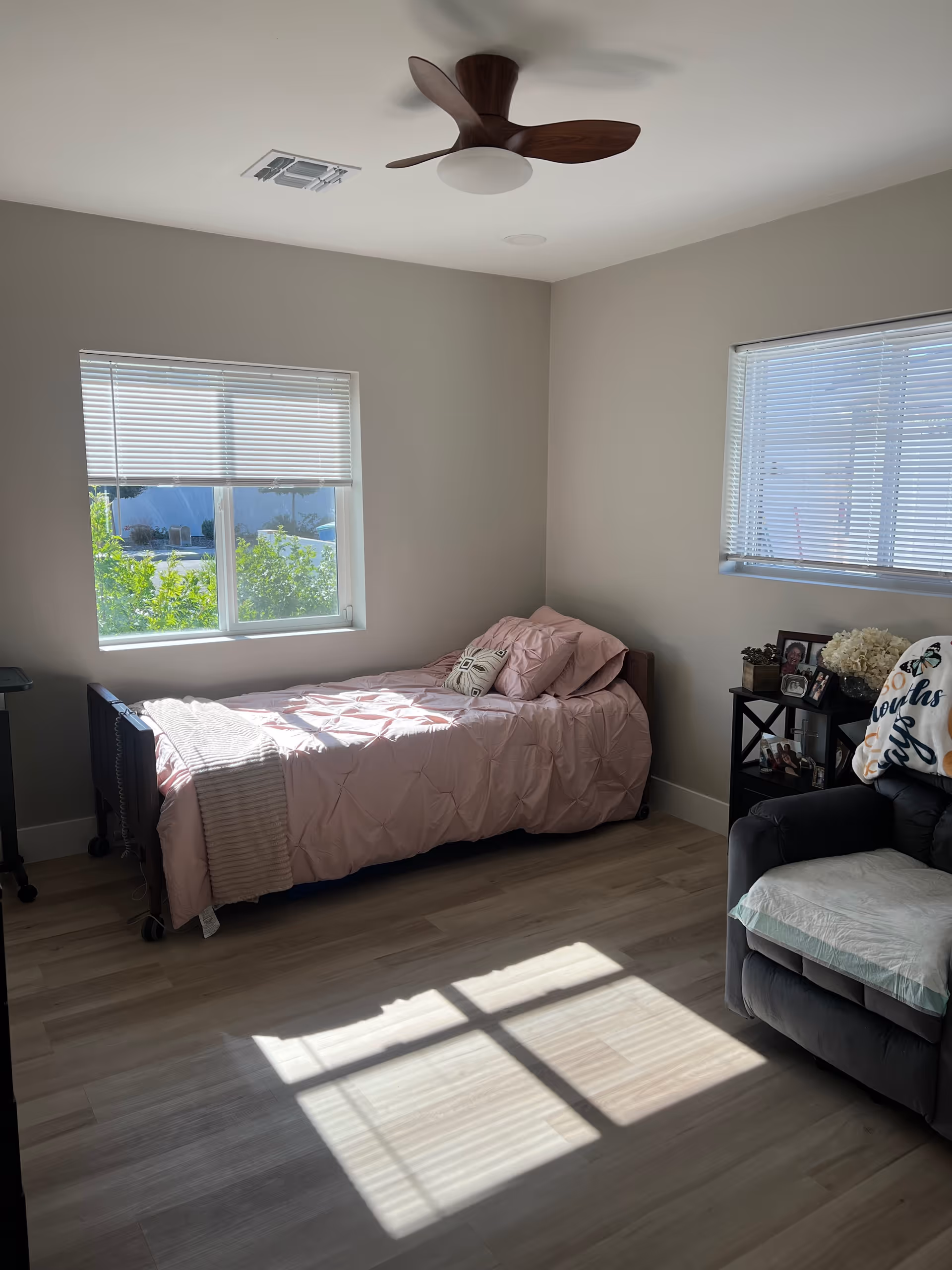 A bright bedroom with two windows letting in natural light. The room features a single bed with pink bedding and pillows, a ceiling fan with wooden blades, a small black side table with framed photos and flowers, and a gray armchair with a white cushion and a decorative pillow.