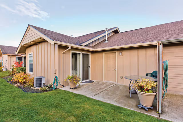 Exterior view of a single-story building with beige siding and a brown shingled roof. There is a sliding glass door and a regular door opening onto a small concrete patio with potted plants and outdoor furniture. The area is surrounded by green grass and some landscaping with small bushes and plants.
