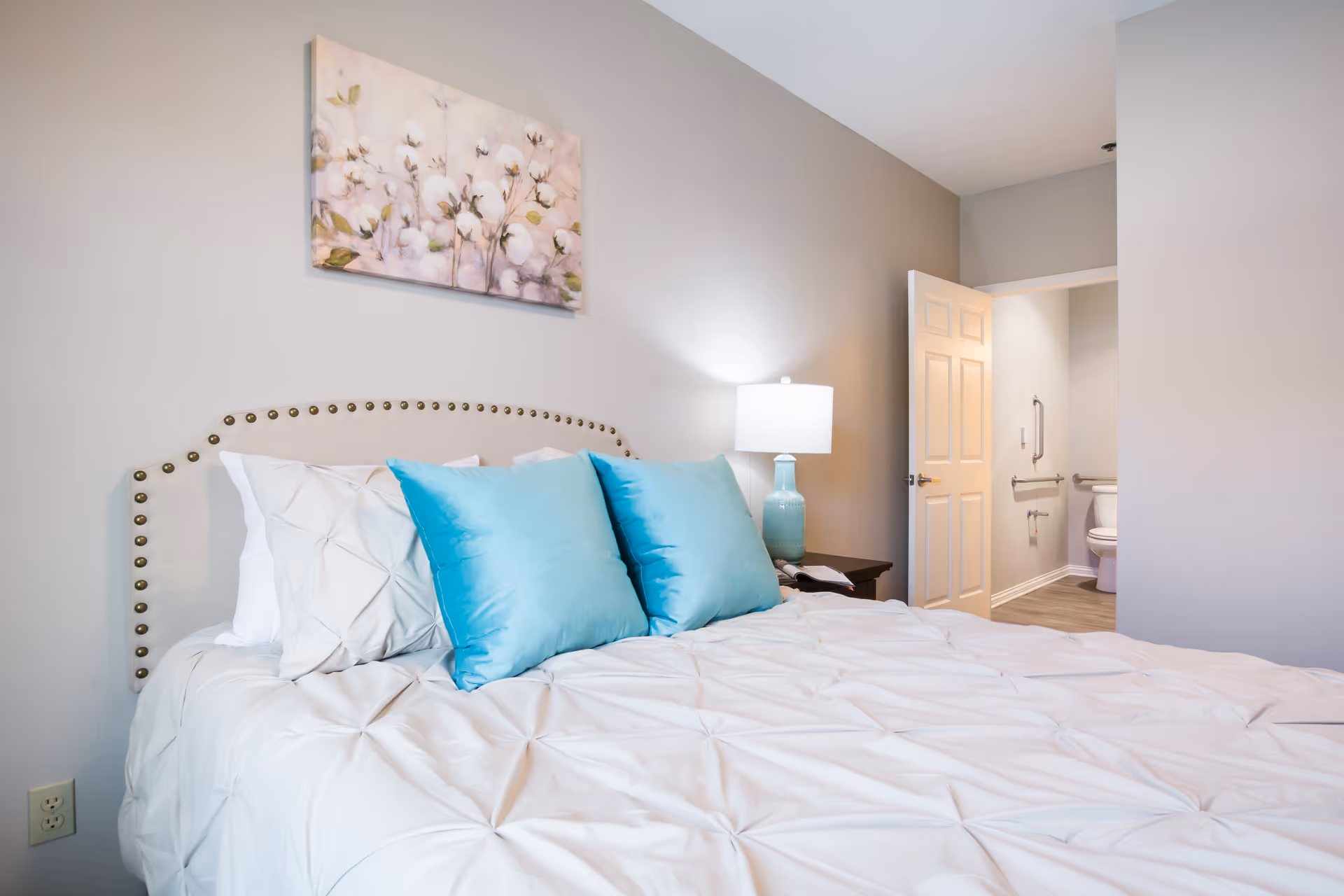 A neatly made bed with white bedding and two turquoise pillows in a bedroom. A nightstand with a turquoise lamp and an open book is beside the bed. A door is open, revealing a bathroom with a toilet and grab bars. A floral painting hangs on the light gray wall above the bed.