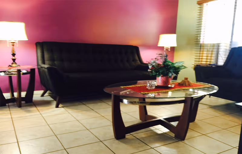 A cozy living room with a dark tufted sofa and armchair, a glass coffee table with a red runner and a potted plant, two side tables each with a lamp, and a purple accent wall in the background.