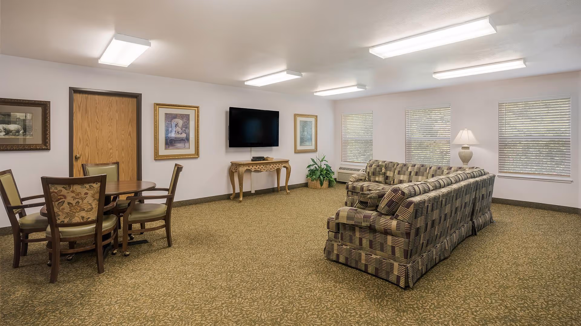 A spacious living room with patterned carpet, a large patterned sectional sofa, a wooden table with four chairs, a flat-screen TV mounted on the wall above an ornate wooden console table, three windows with blinds, a floor lamp, and framed artwork on the walls.