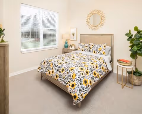 Sunlit bedroom with a bed covered in yellow floral bedding, nightstands, a decorative mirror and a potted plant.