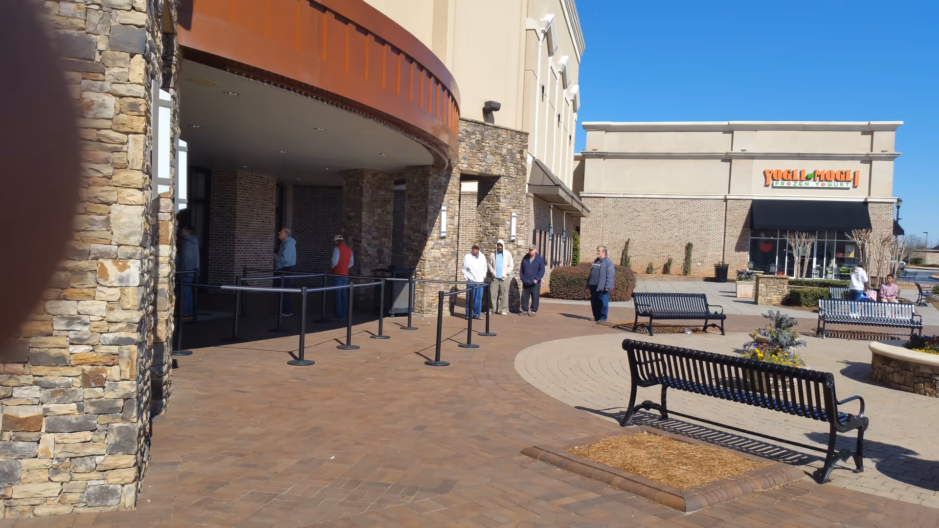 Outdoor shopping area with stone and brick buildings, several people standing and walking near the entrance, black metal benches, and a clear blue sky. A store named 'Yogli Mogli Frozen Yogurt' is visible in the background.