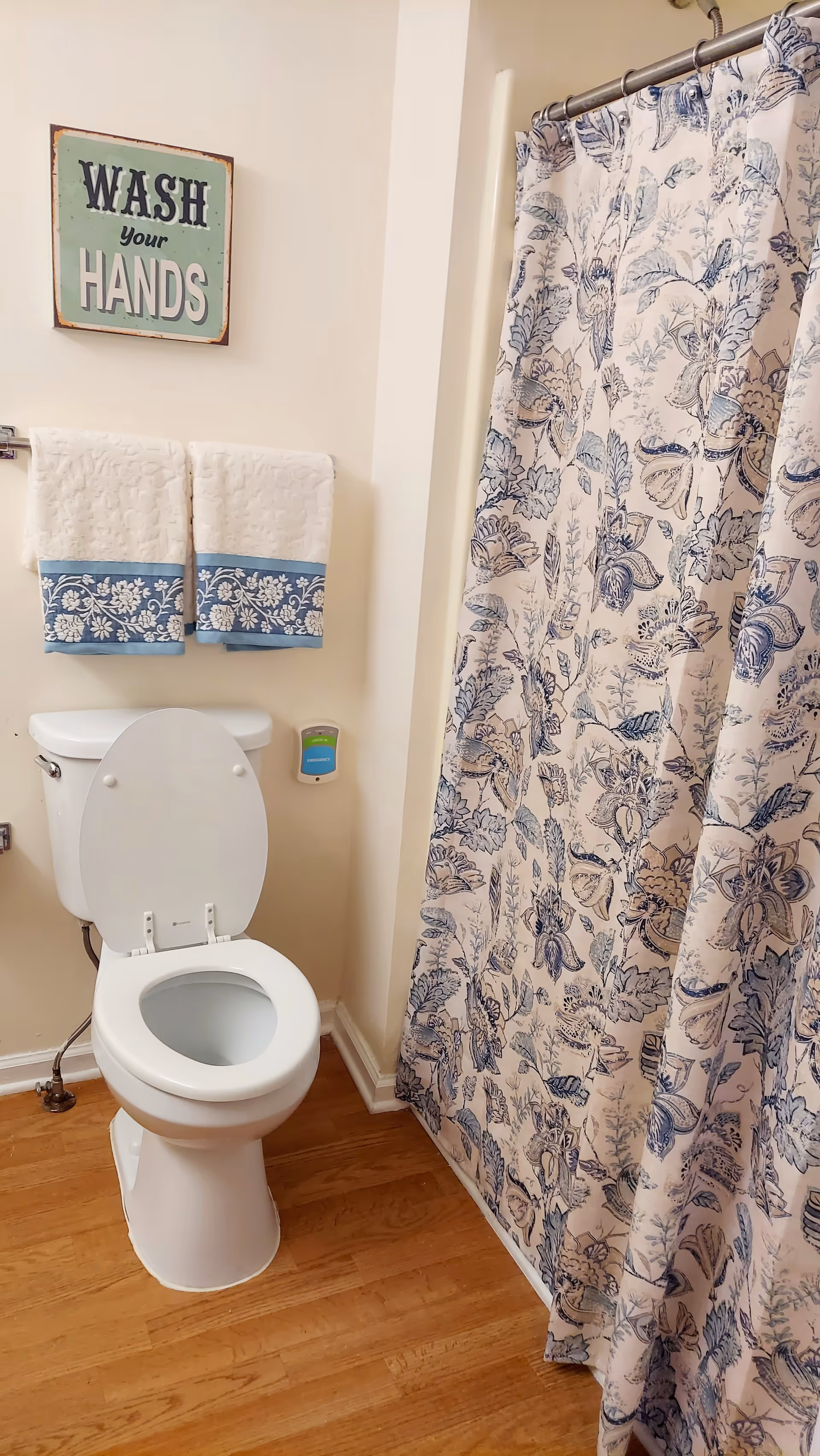 Small bathroom with a toilet, floral shower curtain, hand towels, and a wash-your-hands sign.