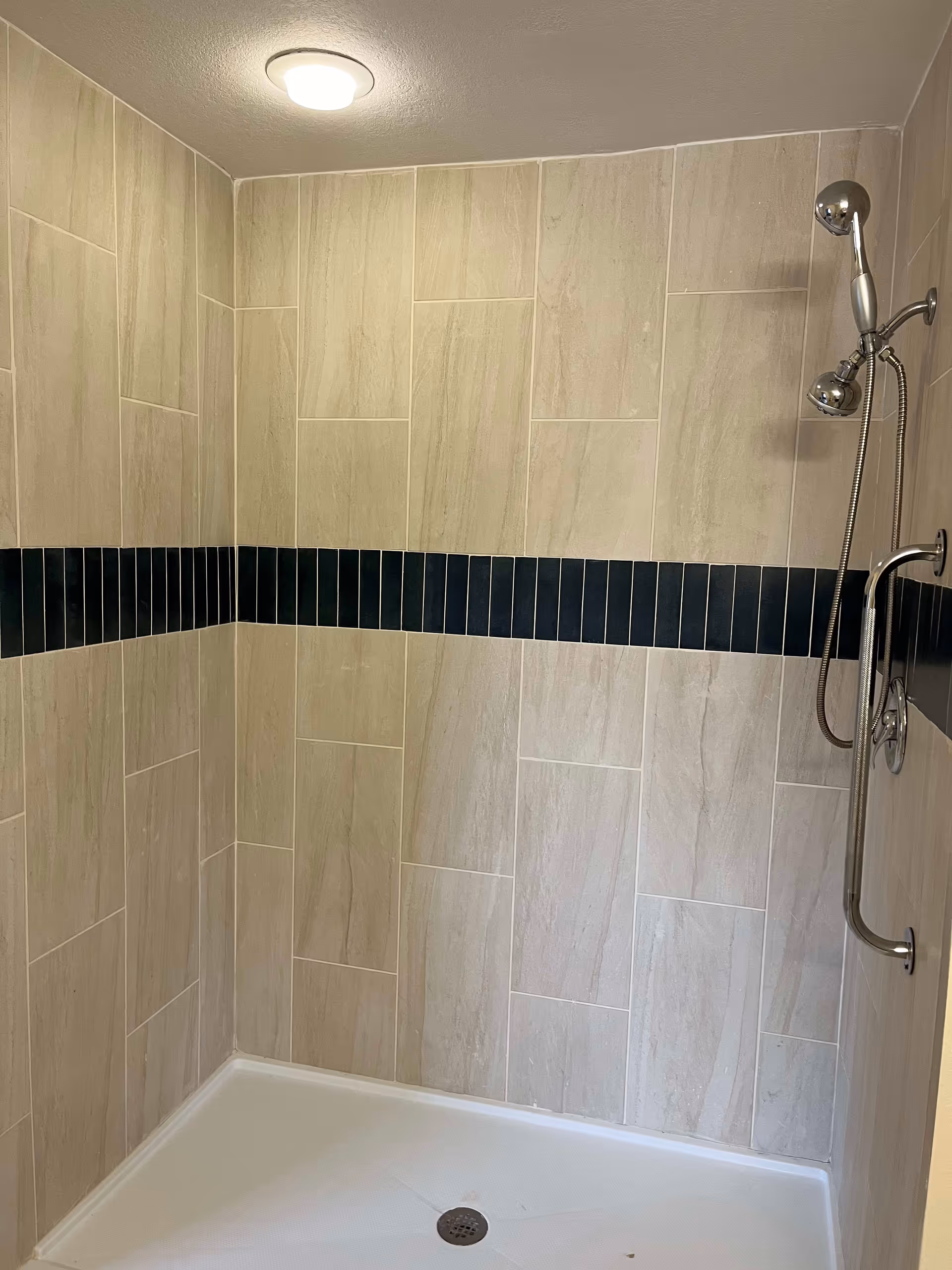 A tiled shower area with beige rectangular tiles and a horizontal strip of dark blue vertical tiles. The shower has a silver handheld showerhead attached to a vertical grab bar on the right wall. The shower floor is white with a central drain.