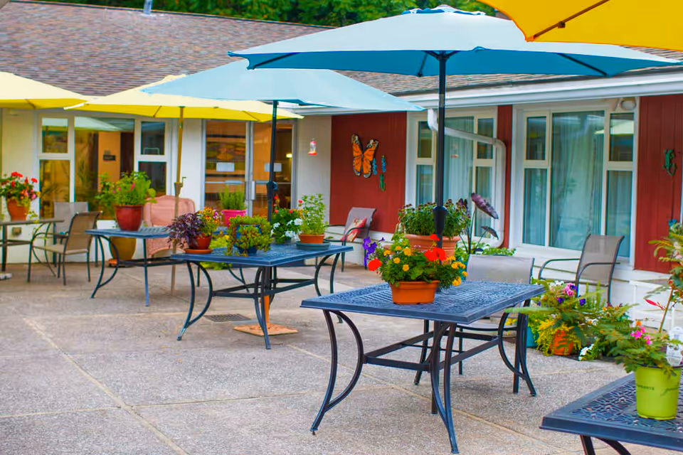 Outdoor patio area with several metal tables and chairs, each table adorned with colorful potted flowers. Large umbrellas provide shade over the tables. The background shows a building with windows, a door, and decorative butterfly wall art.