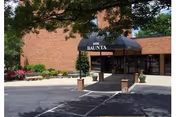 Entrance to a building with a black awning labeled '3216 BAJUNTA' supported by brick pillars, surrounded by a paved driveway and some greenery including bushes and a large tree providing shade.