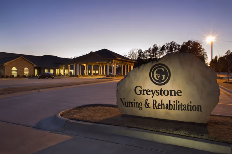 Stone entrance sign reading "Greystone Nursing & Rehabilitation" in front of a nursing facility building at dusk.