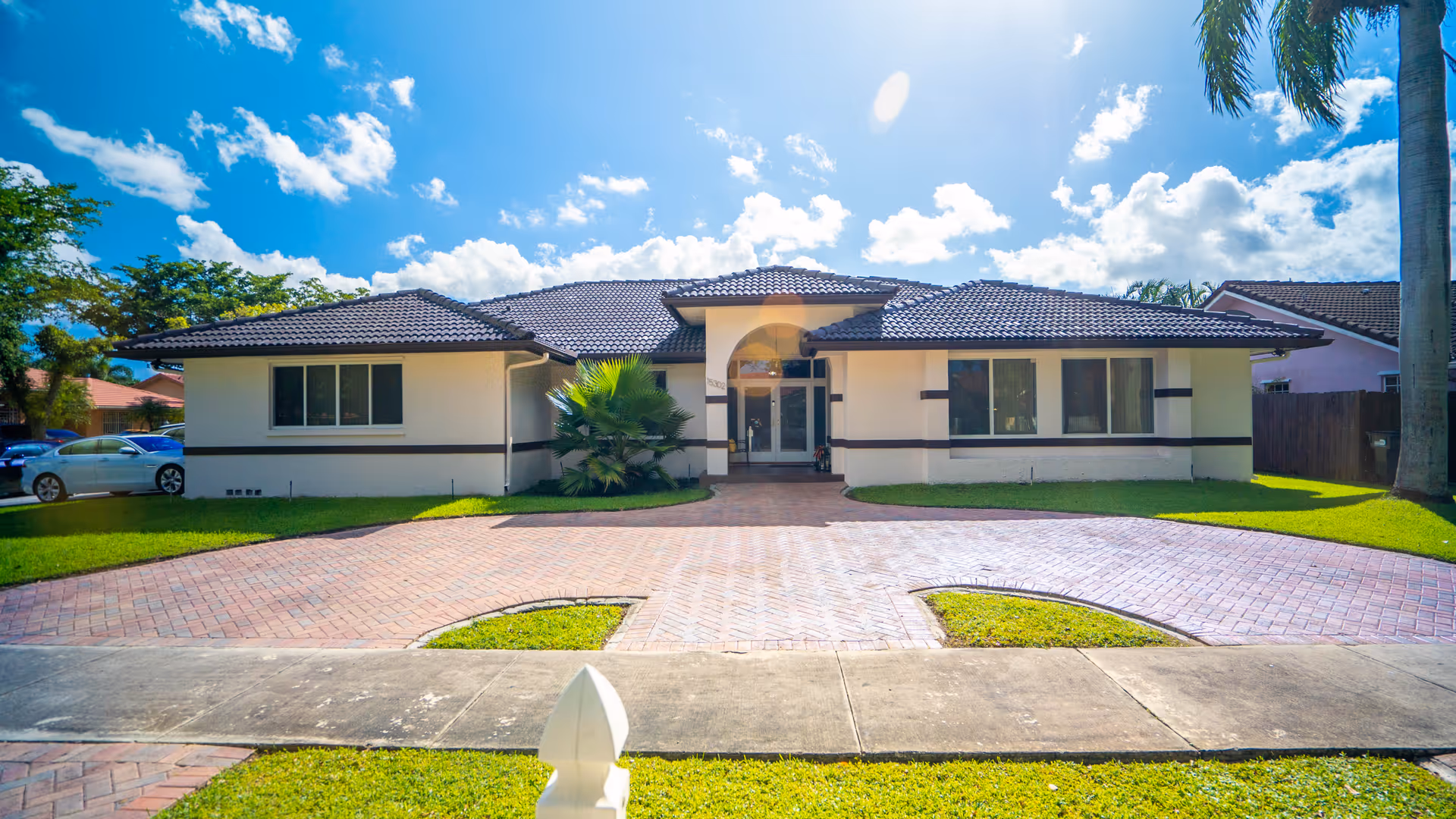 Front exterior view of a single-story building with a tiled roof, large windows, and a paved driveway leading to the entrance. The sky is blue with scattered clouds, and there are palm trees and green grass surrounding the building.