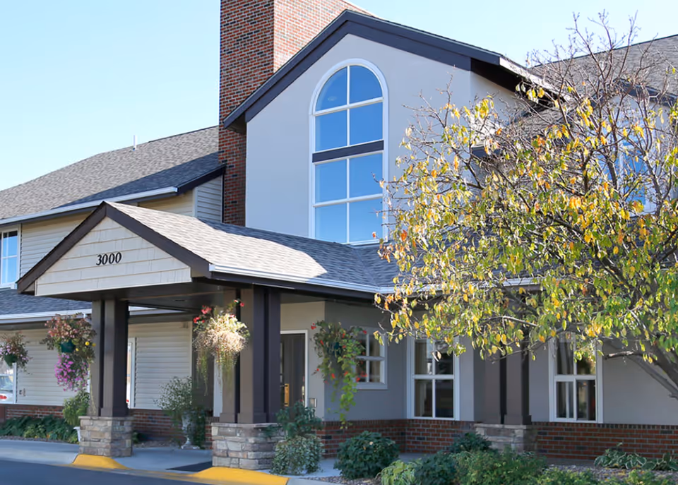 Front entrance of a senior living facility with a covered portico marked "3000", hanging flower baskets, and a tree with autumn leaves.