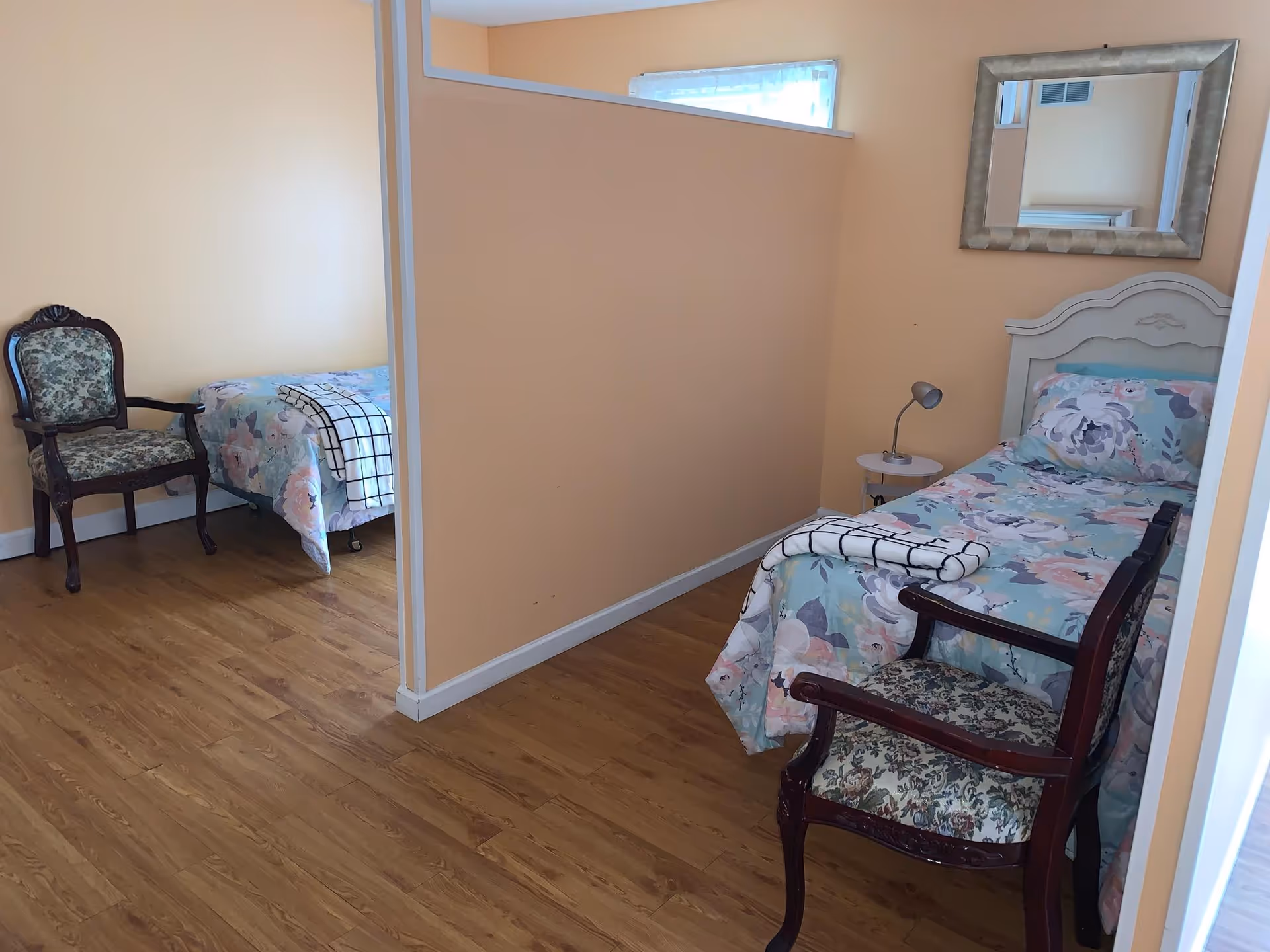 A shared bedroom with two single beds separated by a partial wall. Each bed has floral bedding and a folded blanket at the foot. There is a vintage-style upholstered chair next to each bed. One bed has a small round side table with a lamp and a large decorative mirror on the wall above the headboard. The room has light peach-colored walls and wood flooring.