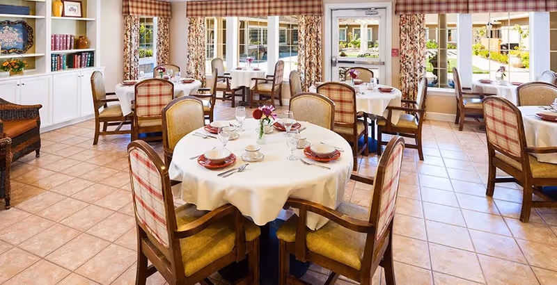 A bright and inviting dining room with round tables covered in white tablecloths, set with plates, bowls, cups, and silverware. The chairs have cushioned seats and backs with a plaid pattern. Large windows with floral curtains and a glass door provide a view of an outdoor courtyard. Shelves with books and decorative items are visible on the left side.