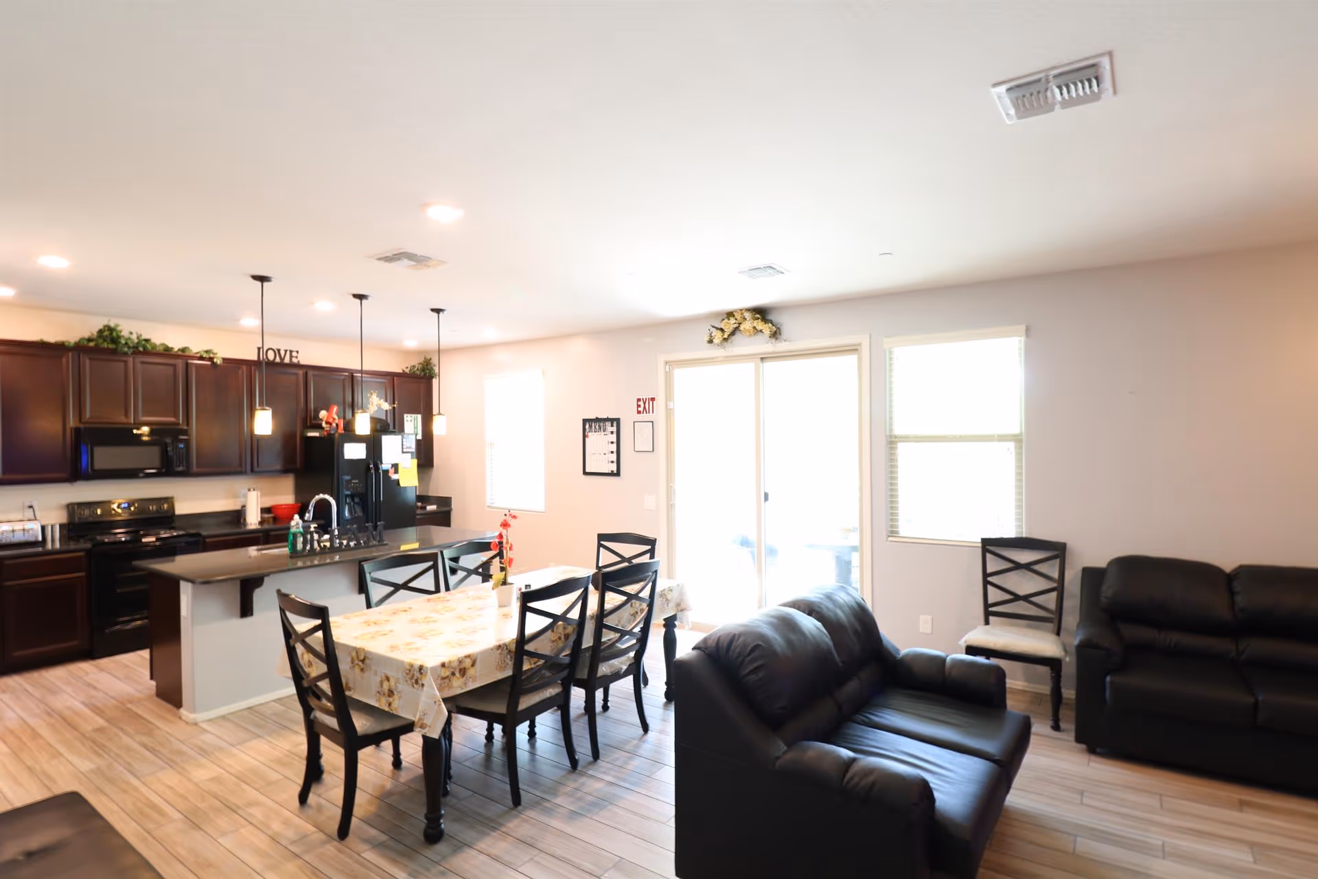 A bright, open interior space featuring a kitchen with dark wood cabinets, black appliances, and a large island with a sink and three pendant lights. Adjacent to the kitchen is a dining area with a table covered by a floral tablecloth and six chairs. The living area includes two black leather sofas and two additional chairs near windows and a sliding glass door leading outside.