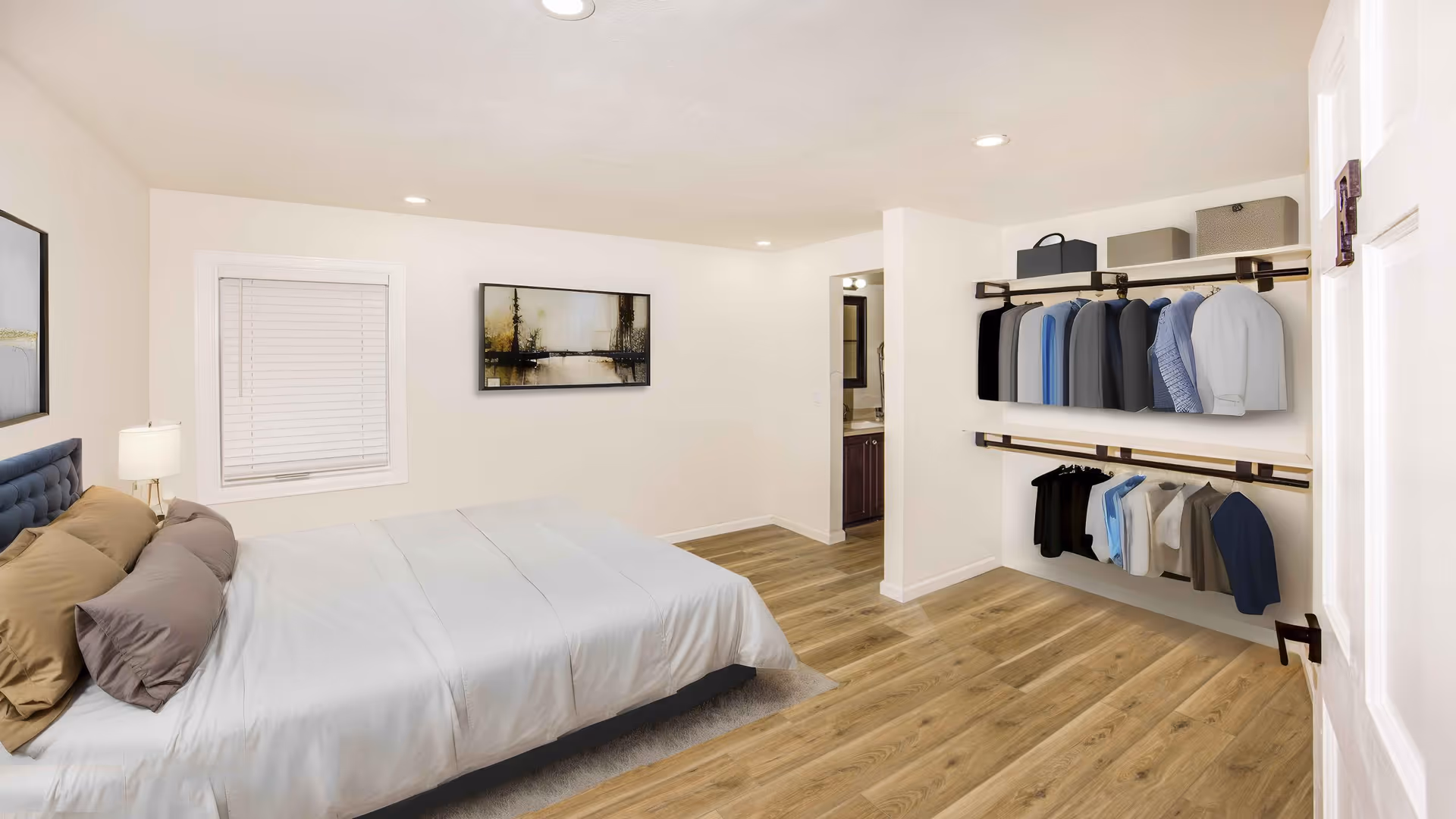 A bright and clean bedroom with a large bed made with white linens and multiple pillows. There is a window with closed blinds on the left wall, a framed landscape painting above the bed, and an open closet on the right side with clothes hanging on two shelves. The room has wooden flooring and recessed ceiling lights. A doorway leads to a bathroom area in the back.