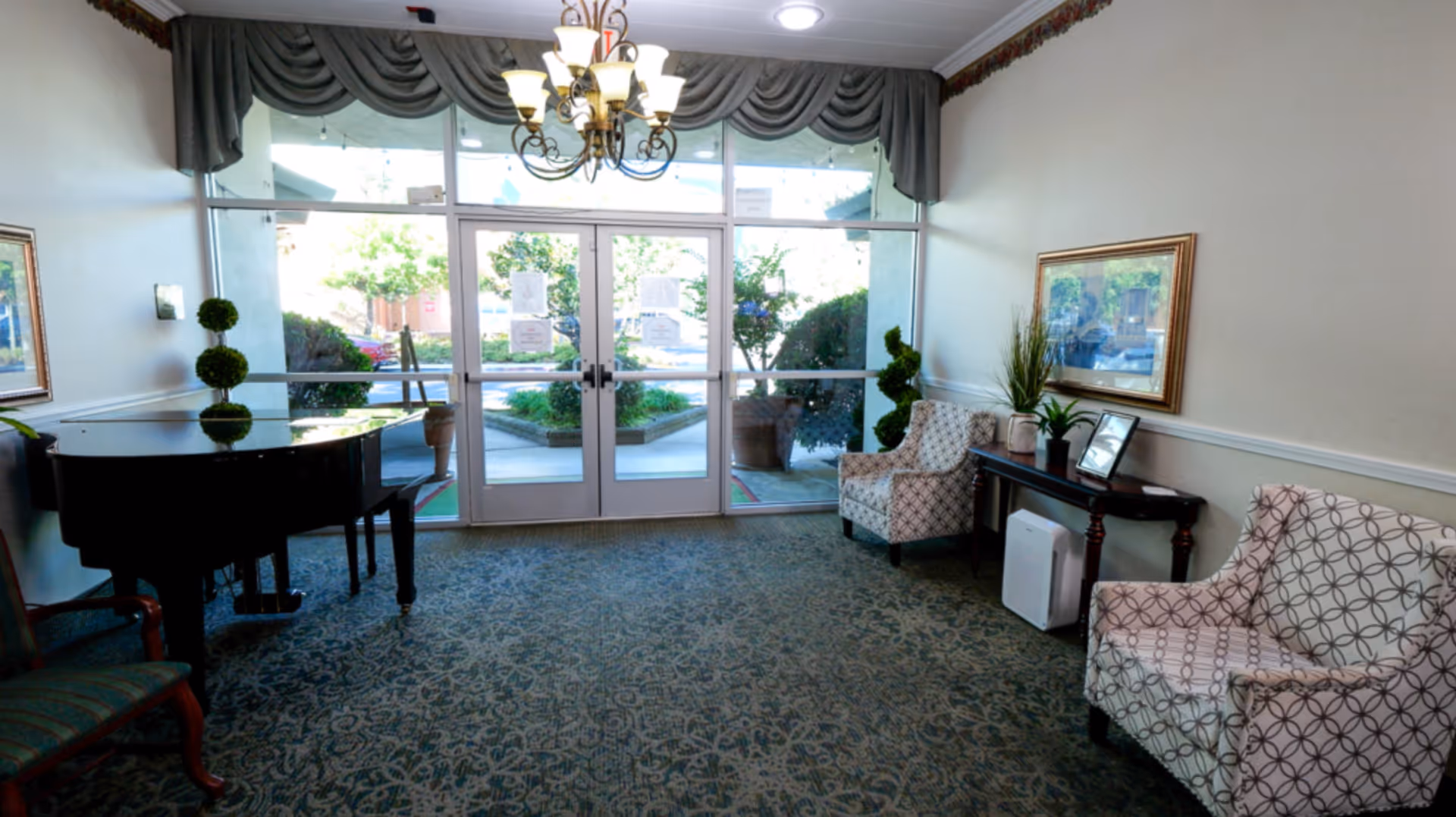 A bright and welcoming lobby area with large glass double doors leading outside. The room features a chandelier, patterned carpet, two upholstered armchairs with geometric designs, a black grand piano, a wooden console table with plants and framed pictures, and a framed painting on the wall.
