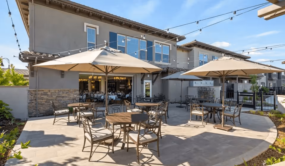 Outdoor patio area at Laurel Heights Senior Living & Memory Care with round tables and chairs under large beige umbrellas. String lights are hung overhead, and the building with large windows is visible in the background under a clear blue sky.