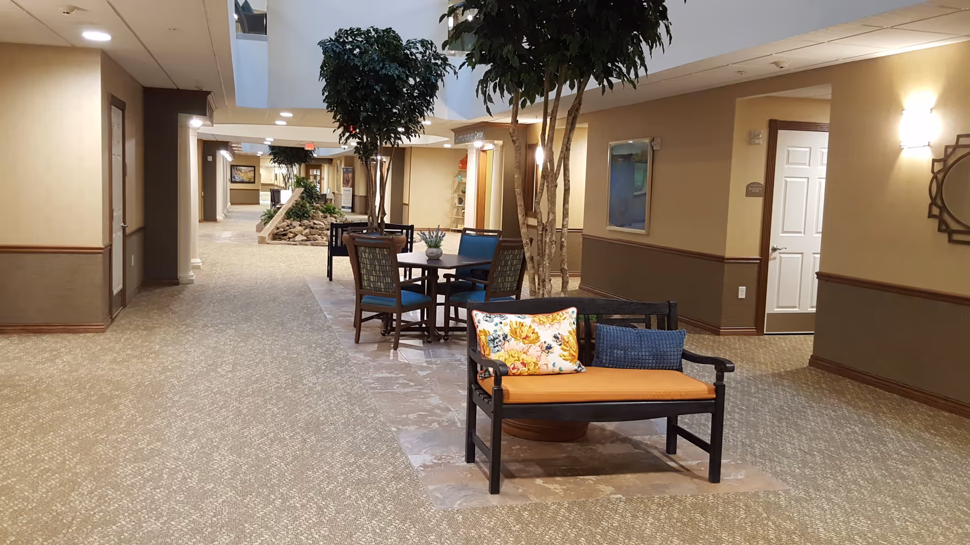 Interior hallway of a retirement community with beige walls and carpeted floors. The hallway features a seating area with a bench that has orange and blue cushions, a table with chairs, and several tall indoor plants. Doors and wall decorations are visible along the corridor, with soft lighting from wall sconces and ceiling lights.