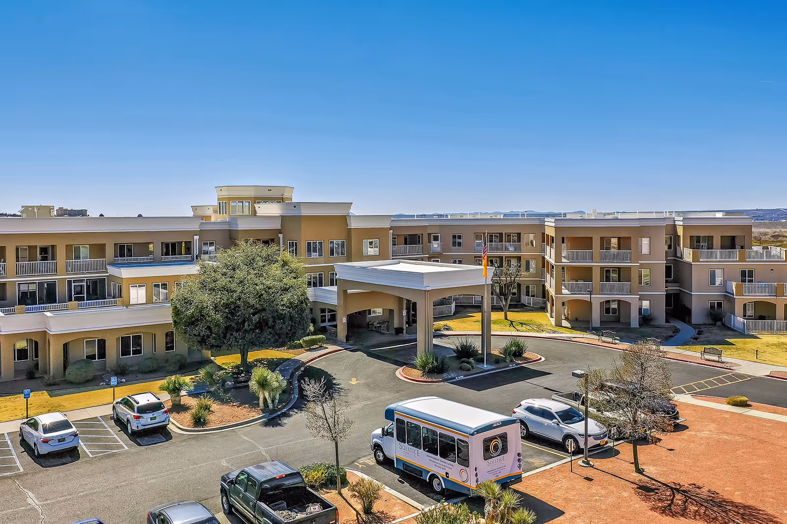 Exterior view of Solstice Senior Living at Las Cruces, showing a three-story building with balconies, a covered entrance, several parked cars, and a shuttle bus in the parking lot under a clear blue sky.