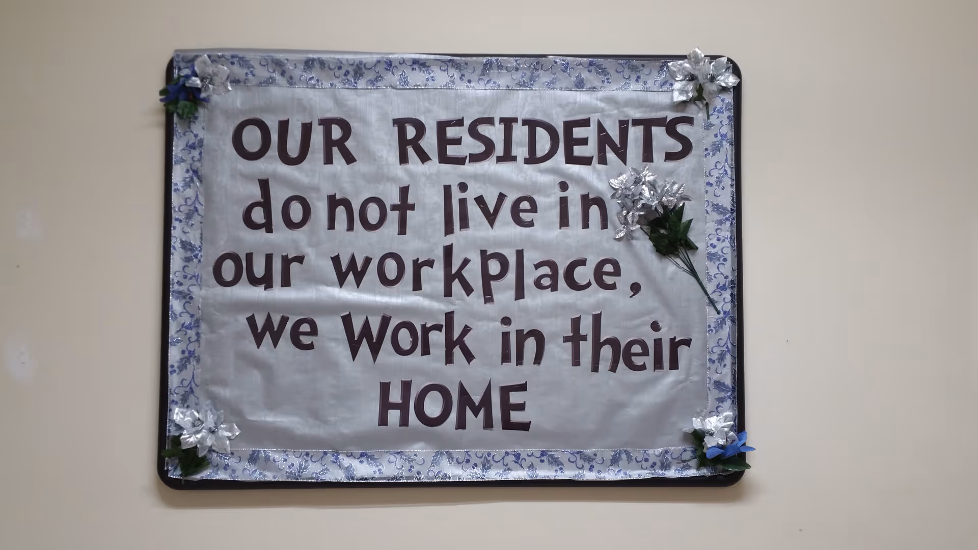A decorative bulletin board with a silver background and floral border displaying the message: 'OUR RESIDENTS do not live in our workplace, we Work in their HOME' with silver and blue flower decorations in the corners.