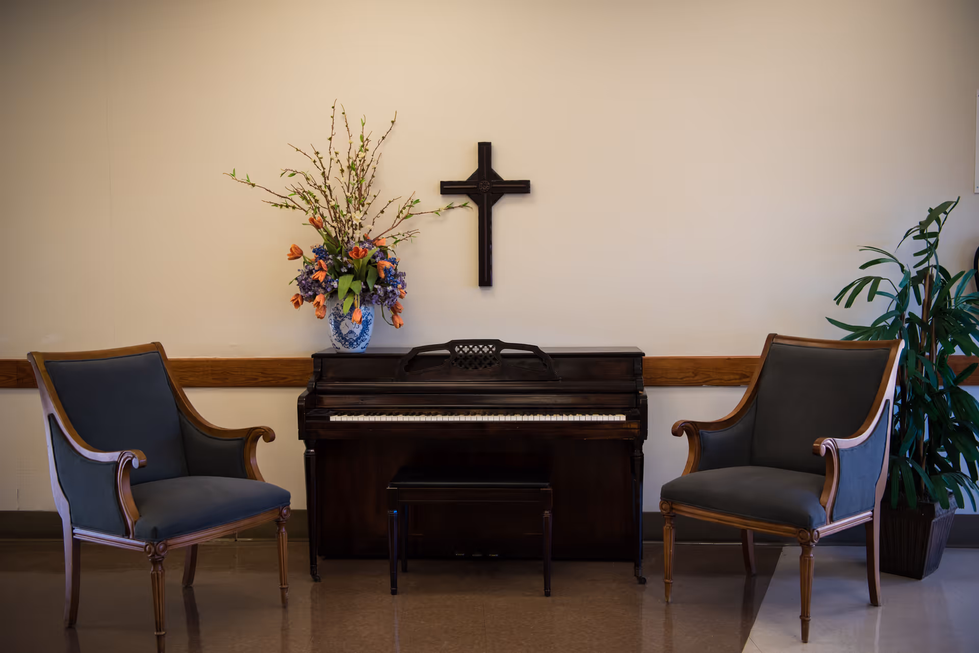 Upright piano flanked by two upholstered armchairs with a vase of flowers and a cross on the wall above.