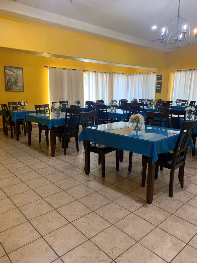 A dining room with multiple tables covered in blue tablecloths, each table surrounded by dark wooden chairs. Each table has a small vase with white flowers. The room has yellow walls, tiled floors, large windows with white curtains, and a chandelier hanging from the ceiling.