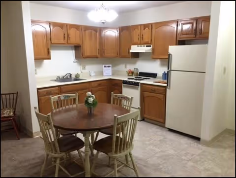 A kitchen with wooden cabinets, a white refrigerator, a stove with a range hood, a sink, and a round wooden dining table with four chairs. A small flower arrangement is placed in the center of the table.