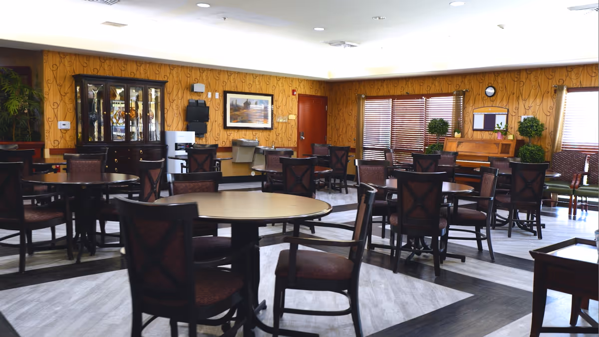 A spacious dining room with multiple round tables and chairs arranged neatly. The walls have a warm yellow patterned wallpaper, and there is a wooden cabinet with glass doors displaying decorative items. A piano is placed against the far wall with plants and framed pictures around it. Large windows with blinds allow natural light to enter the room.
