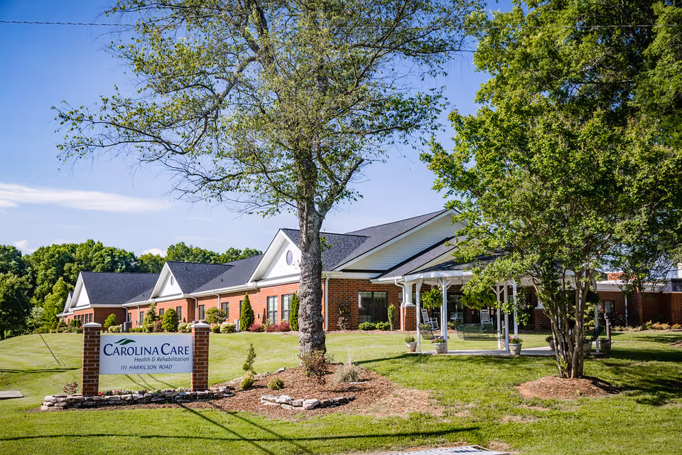 Exterior view of Carolina Care Health and Rehabilitation with a brick front, landscaped lawn, trees, and a sign at the entrance.