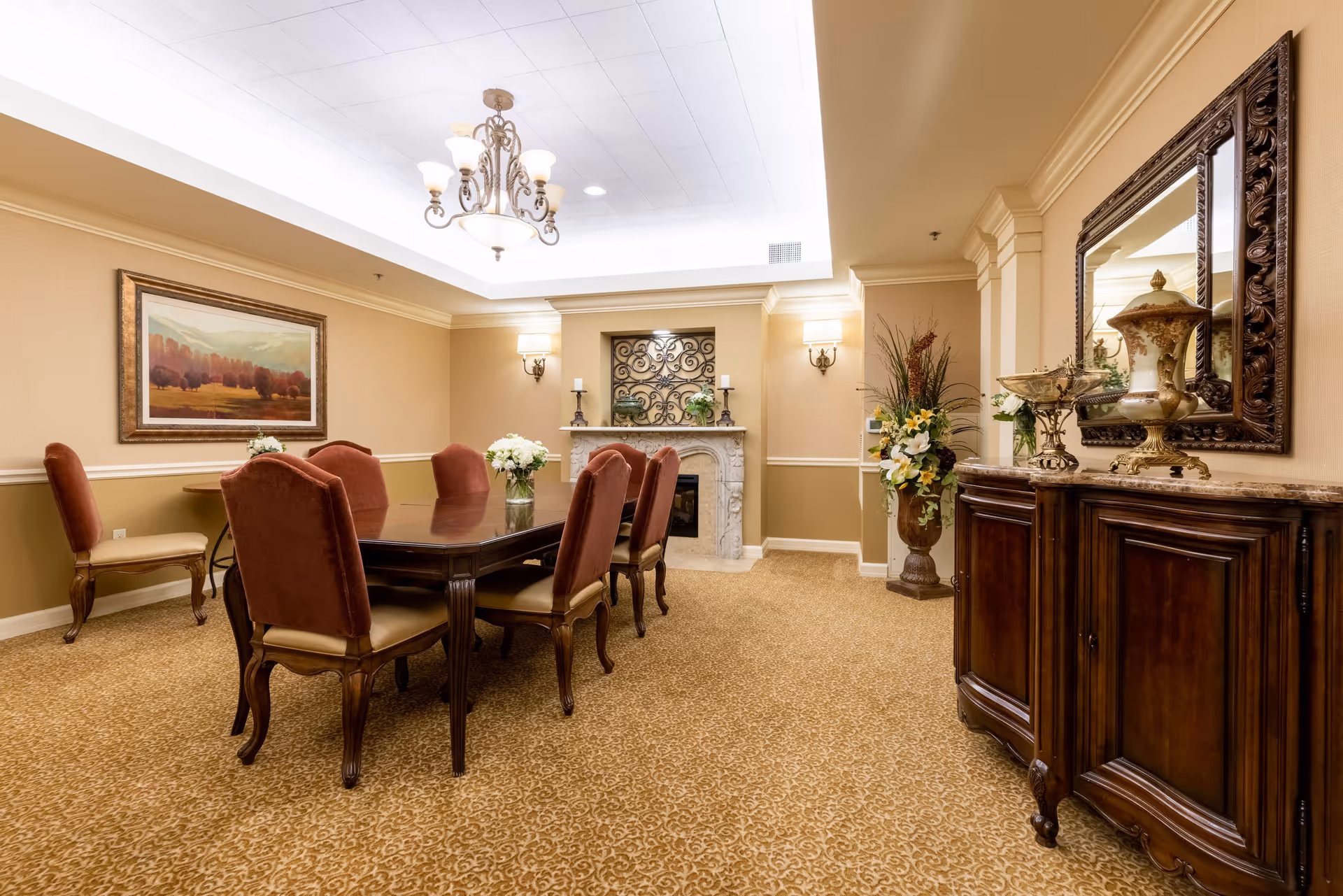 A formal dining room with a long wooden table surrounded by eight upholstered chairs. The room features a beige and cream color scheme with patterned carpet, a decorative fireplace with candles and plants, a large framed painting on the wall, a chandelier hanging from the ceiling, and a wooden sideboard with ornate vases and a large mirror above it.