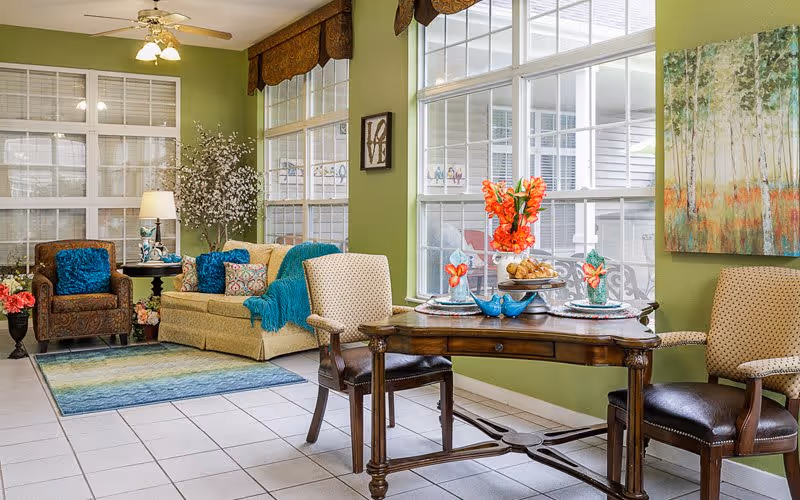 A bright and cozy living room area with green walls, large windows, and white tiled floor. The room features a beige sofa with blue and patterned cushions, a brown armchair, a wooden table set for two with floral decorations, and a colorful area rug. There are decorative plants, a ceiling fan with lights, and framed artwork on the walls.