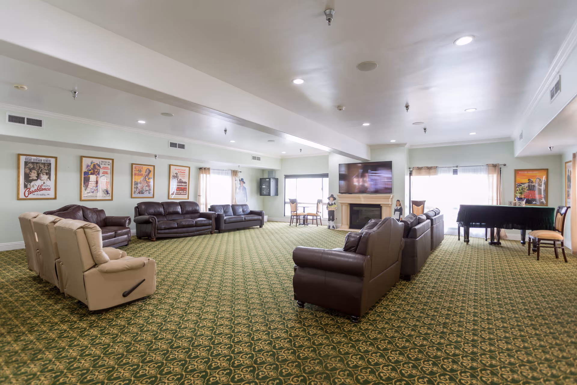 A spacious living room area with green patterned carpet, multiple brown leather sofas, and beige recliners arranged around a large flat-screen TV mounted above a fireplace. The walls are decorated with framed vintage movie posters, and there is a black grand piano with chairs near large windows with curtains letting in natural light.
