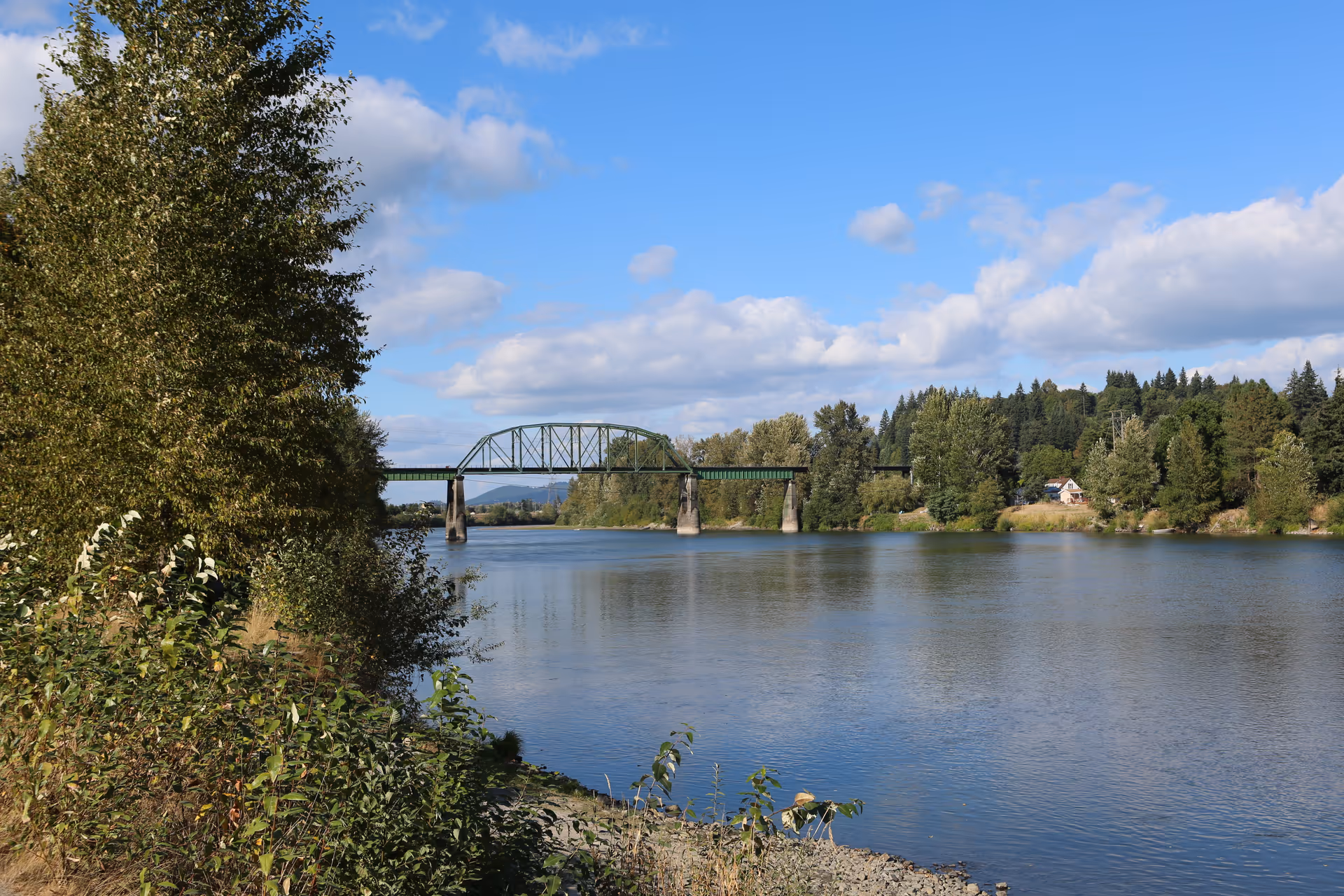 A scenic view of a river with a green metal bridge crossing over it. The riverbank is lined with trees and bushes, and there are more trees and a few houses visible on the opposite side of the river under a partly cloudy blue sky.
