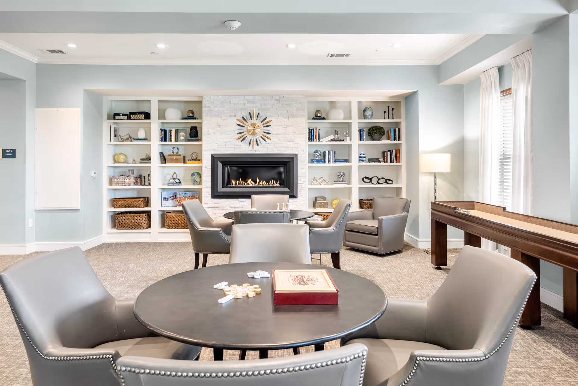 A cozy living room area with a modern fireplace centered on a white stone wall flanked by built-in shelves filled with books and decorative items. In front of the fireplace are two round tables with gray leather chairs around them. A shuffleboard table is positioned near a window with white curtains, and a floor lamp stands in the corner providing soft lighting.