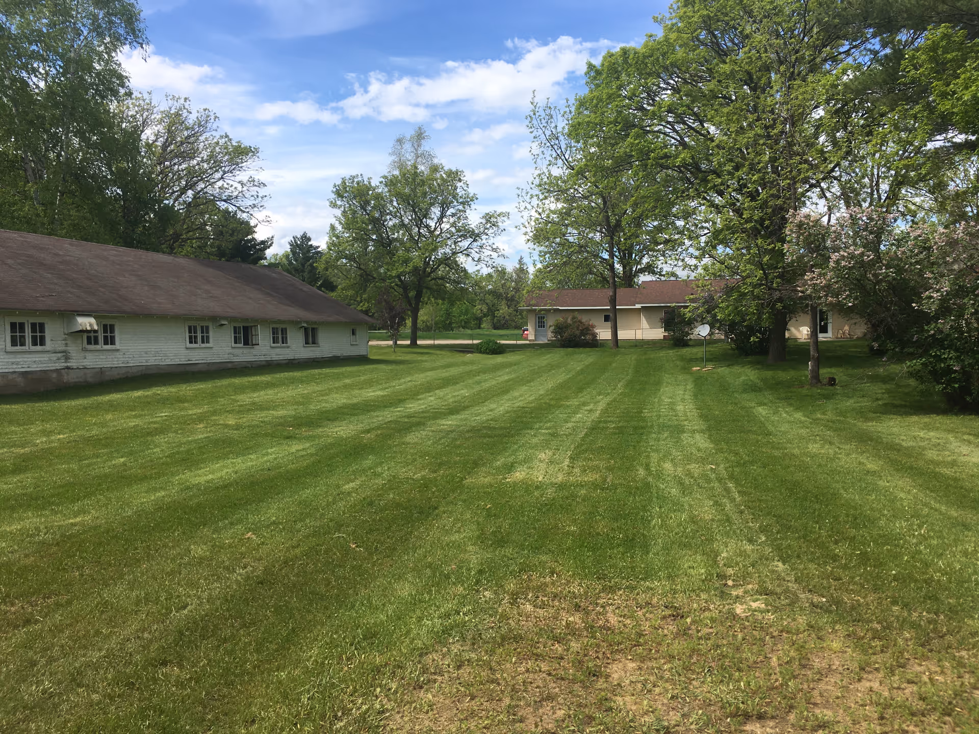 Mowed grassy lawn flanked by single-story buildings and trees under a partly cloudy sky.