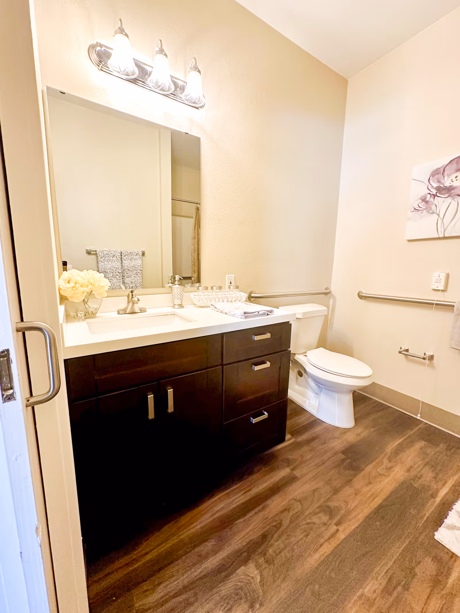 Well-lit bathroom with a dark wood vanity and sink under a mirror, a toilet with grab bars, and wood-look flooring.